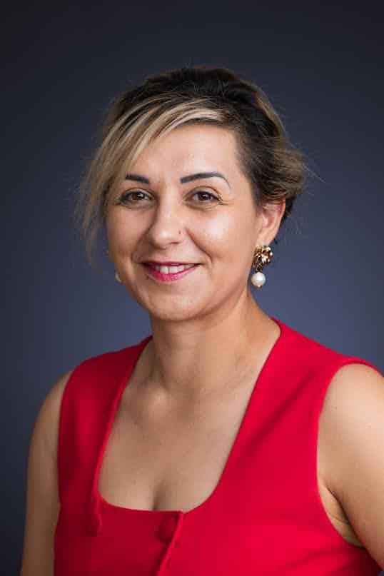 Femme souriante en haut rouge et boucles d'oreilles perles, cheveux méchés blonds, piercing nez.