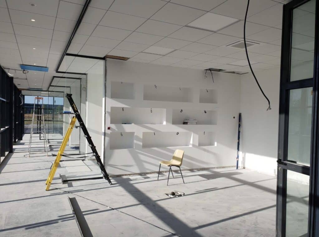 Chantier intérieur lumineux avec murs blancs, niches encastrées, échelle jaune et noire, sol en béton brut, et câbles pendant.